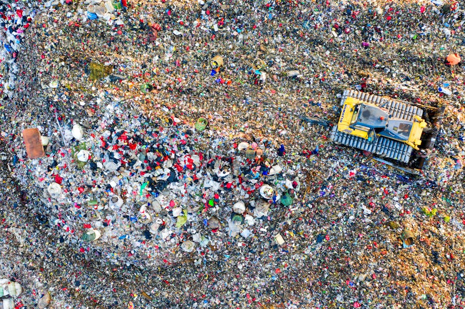An aerial view of a section of a large landfill site filled with a dense layer of mixed waste debris, including plastic bottles, containers, paper, and other household refuse in various colors such as red, white, blue, and green. The ground surface beneath the waste appears uneven and compacted, with visible mounds and strips of dirt, indicating ongoing waste material layering. On the right side of the image, a yellow tracked excavator with a metal bucket attachment is actively moving or sorting rubbish, situated on a strip of exposed earth that appears clearer of debris, suggesting recent or ongoing waste removal activity. The surrounding environment shows a stark contrast between the heavily cluttered waste area and the exposed earth sections, highlighting the scale of waste accumulation typical of unmanaged or inadequately managed waste sites. This scene underscores the importance of professional rubbish clearance services like those provided by wasteclearancewestkensington.co.uk to prevent illegal dumping and promote environmentally responsible waste disposal, emphasizing the process of on-site clearance that assists in managing waste effectively and avoiding fines from local authorities for illegal rubbish dumping.