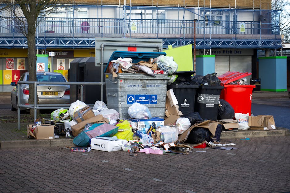 A large open area covered with a mixture of waste materials, predominantly black rubber tires that vary in size and are scattered across the foreground. Interspersed among the tires are white and colored plastic bags, cardboard boxes, and miscellaneous household rubbish. The waste appears unorganized and extends across the entire width of the image, with some debris piled haphazardly. In the background, there is a natural landscape with green, densely wooded hills under a partly cloudy sky, contrasting with the foreground of discarded rubbish. This scene exemplifies an unmanaged or illegal waste disposal site, highlighting the importance of professional rubbish removal services like those offered by waste clearance specialists to prevent environmental pollution and support proper waste handling.