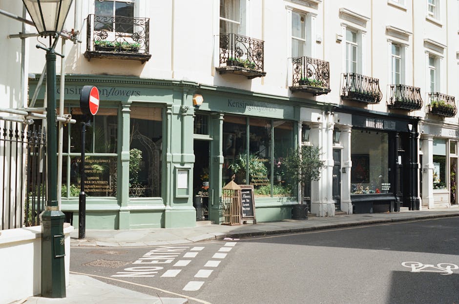 The image depicts a row of commercial storefronts on a street corner, with the corner store painted in light green featuring large glass windows displaying plants and flowers inside. A black signboard stands outside the store on the pavement, possibly advertising products or services. Adjacent to it are other shopfronts with white and black facades, also with large display windows containing various items. Above the shops, there are several residential-style balconies with ornate black wrought-iron railings and flower boxes filled with greenery. A lamppost stands at the street corner, near a no-entry traffic sign, with a black metal fence running along the sidewalk. The pavement is clean, with a designated bicycle lane marked on the road surface. The overall scene is well-lit, with natural daylight illuminating the building facades and street, evoking a typical urban setting that may relate to independent retail establishments that could benefit from external waste removal or clearance services offered by companies like wasteclearancewestkensington.co.uk, highlighting the importance of proper rubbish management for commercial premises in a city environment.