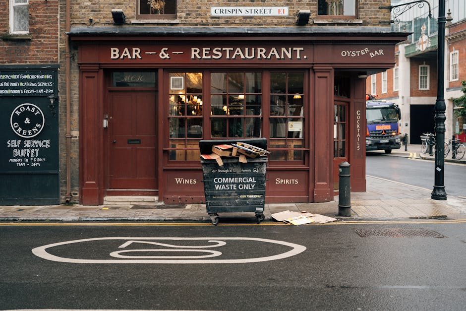 A black waste bin labeled 'Commercial Waste Only' is placed outside a closed restaurant on a city street, with cardboard boxes and paper waste stacked haphazardly on top. The restaurant has a brown wooden façade with large windows, and a sign indicating it is a bar and restaurant specializing in wines and spirits, with an oyster bar section. To the left, a blackboard sign advertises a self-service buffet, and a street sign above reads 'Crispin Street E1'. The bin is positioned on a narrow sidewalk beside a yellow line on the road, which features a white directional arrow painted on asphalt. Nearby, a black bollard and a partly visible black street lamp are present, with a delivery truck parked further down the road. The environment suggests an urban setting, highlighting the importance of proper waste management and alternative collection options for businesses like these, which may occasionally generate waste that needs professional disposal services. The scene emphasizes the potential for improperly managed waste outside commercial establishments and underscores the relevance of dedicated rubbish removal services such as those offered by wasteclearancewestkensington.co.uk for local landlords and business owners.