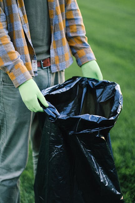 A person wearing a casual plaid shirt with yellow and gray tones, a dark green T-shirt, and gray trousers is standing outdoors on a grassy area during the daytime. They are holding open a large black plastic rubbish bag lined with a silver interior, with their hands gloved in bright green cleaning gloves. The person's face is not visible in the image, and the focus is on their hands and the rubbish bag, suggesting they are in the process of collecting or disposing of waste. The background features lush green grass, indicating an outdoor setting such as a garden, park, or outdoor area. The scene conveys a context of waste management or rubbish collection, possibly related to private or independent waste clearance services like those offered by wasteclearancewestkensington.co.uk, with an emphasis on responsible rubbish removal and disposal.