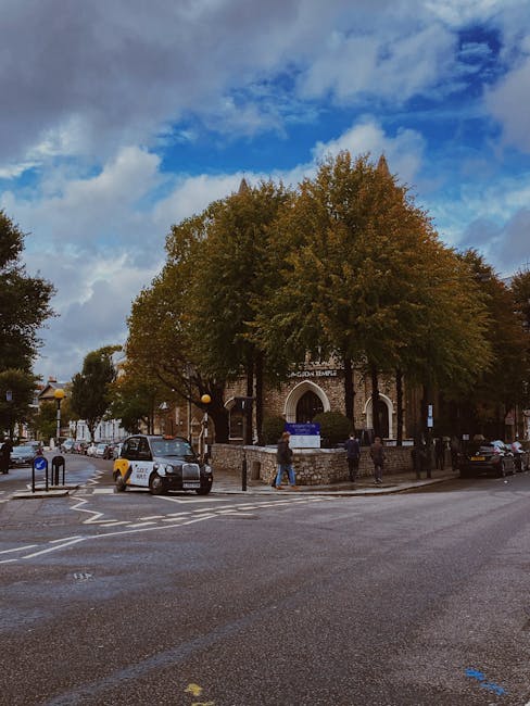 A street scene in a residential or commercial area featuring a large, mature tree with dense, green and brownish leaves situated in front of a brick building with arched entrance doors. The building appears to be a church or community centre, with signage above the entrance. Several parked cars, including a black hatchback and a white vehicle, are visible along the curb, with some in a designated parking zone marked by blue and yellow bollards. The pavement and road surface show signs of recent rain, with wet patches and reflective surfaces, while a few pedestrians walk along the sidewalk or crossing the street. Overhead, a partly cloudy sky with patches of blue peeking through suggests a mix of sun and cloud, creating natural light that highlights the textures of the building's brickwork and the tree’s foliage. This scene captures a typical urban environment where occasional rubbish clearance services might be needed for maintaining cleanliness around public spaces, with the setting reflecting typical West Kensington surroundings served by independent waste collection providers like wasteclearancewestkensington.co.uk.