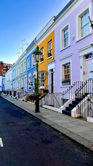 The image shows a row of terraced houses on Fulham Palace Road, each with distinct pastel-colored facades including light blue, yellow, and lavender. The houses are two or three stories tall, featuring white-framed windows and small front doors, some with decorative trim. Black wrought-iron railings line each front step, leading up to the entrance doors. In the foreground, a traditional black lamppost with a green top is positioned on the sidewalk, which is paved with large rectangular stone slabs. The street surface appears dark, smooth asphalt, with faint white street markings. The environment is illuminated by natural daylight, casting soft shadows along the pavement and building facades. Visible on the left side are rooftops with antennas, and in the background, there are additional buildings suggesting an urban residential area. This scene is typical of a well-maintained urban street where private waste collection, such as rubbish clearance services provided by companies like wasteclearancewestkensington.co.uk, may be part of scheduled or on-demand waste removal processes instead of council collection, particularly for bulky waste or special disposal needs.