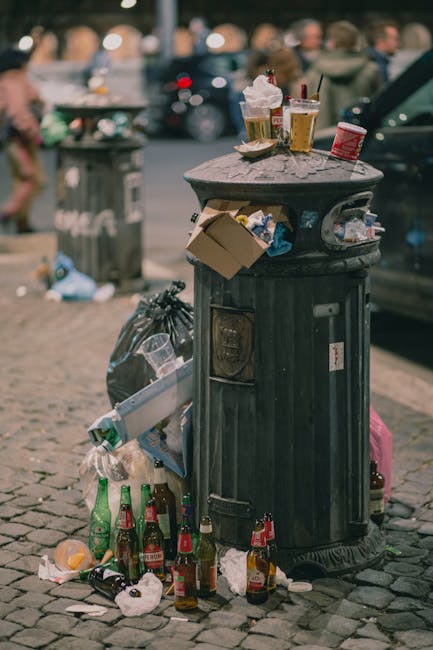A public street scene featuring an overflowing black waste bin with a rounded lid, situated on a cobblestone pavement. The bin is cluttered with discarded cardboard, plastic bottles, and paper cups. Several glass beer bottles are scattered on the ground in front of it, some still partially filled with liquid, along with crumpled paper and other small debris. On top of the bin, two cups with remnants of drinks and plastic straws, as well as a cardboard container, are visibly placed. In the background, there is a blurred view of parked cars and a green trash bag leaning against the bin, indicating abandoned rubbish. The overall scene reflects littering and improper waste disposal typical of urban environments, relevant to private waste management or rubbish removal services. The natural lighting highlights the texture of the cobblestones and the weathered surface of the bin, emphasizing the importance of effective waste clearance to maintain street cleanliness.