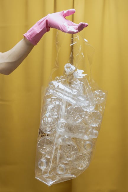 A person wearing a light pink rubber glove is holding a clear plastic bag filled with discarded plastic bottles and containers. The bag is partially crumpled, with the bottles inside appearing transparent and of various shapes and sizes, some with caps and others without. The background consists of a plain yellow curtain or fabric, providing a neutral backdrop that highlights the waste materials. The scene emphasizes the importance of proper waste collection and disposal, relevant to private rubbish clearance services, such as those offered by waste clearance companies. The image visually represents the process of sorting and collecting recyclable plastic waste, which may be part of an on-site rubbish clearance carried out independently or through alternative waste handling methods outside local authority collection systems.