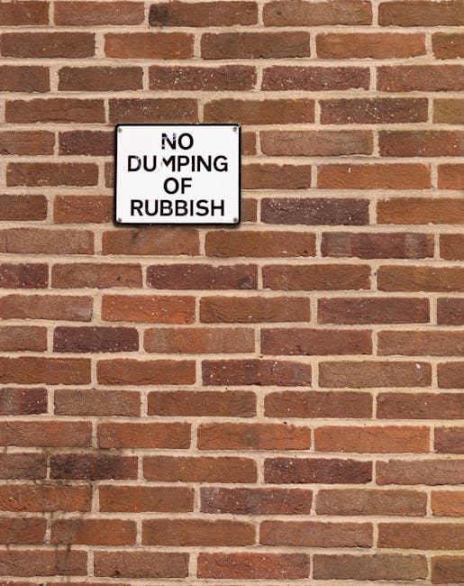 A white rectangular sign with black bold lettering mounted on a brick wall, displaying the message 'NO DUMPING OF RUBBISH'. The brick wall behind the sign is composed of reddish-brown bricks with a slightly textured surface, arranged in a traditional horizontal pattern with light-colored mortar joints. The sign is positioned toward the upper left of the image, and the wall extends uniformly across the background. The lighting appears natural, with no significant shadows on the wall or sign. This visual context relates to waste management and the importance of proper rubbish disposal, aligning with services such as private rubbish removal or site clearance handled by waste clearance specialists like wasteclearancewestkensington.co.uk.