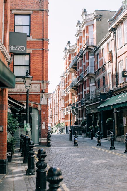 A narrow urban street with a cobblestone surface, lined by multi-storey red and beige brick buildings with decorative balconies and large windows. On the left, there is a shop sign reading 'HEZZO' attached to a brick building, with a traditional black street lamp mounted nearby. The right side features storefronts under green awnings, with small hanging plants and outdoor seating areas. In the foreground, black bollards separate the pedestrian walkway from the street, and a few potted plants are visible near the shops. The street is illuminated by natural daylight, creating a bright and clear environment suited for pedestrian access. The scene reflects a typical city centre atmosphere with a mix of commercial and residential architecture, where independent shops and local businesses may utilize private waste collection services like those provided by companies such as wasteclearancewestkensington.co.uk, for effective rubbish removal and on-site clearance in a densely built environment. The overall setting suggests a well-maintained area supporting sustainable waste disposal options for retail and residential properties alike.