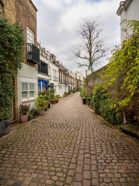 A narrow residential cobblestone street lined with a variety of terraced houses featuring multiple stories, white and pastel-colored facades, and small balconies with metal railings. Potted plants and small shrubs are positioned along the base of the buildings, adding greenery to the scene. On the right side, a high brick wall is partially covered with green climbing plants and foliage, with some plants spilling over the top. In the background, a large leafless tree with numerous thin branches stands prominently against an overcast sky, casting diffuse natural light across the scene. Black wheelie bins are placed along the street, indicating waste collection points, and the overall environment reflects a quiet, urban residential area with a historic European character. Wasteclearancewestkensington.co.uk sometimes handles private waste and on-site clearance, which is visually supported by the presence of waste bins in this setting, emphasizing alternative waste handling outside of local authority collections.