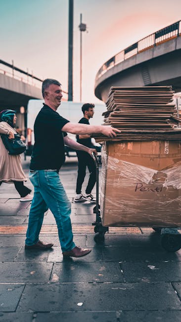 A man in a black long-sleeved shirt and blue jeans is pushing a large, open-top wheeled collection bin filled with flattened cardboard boxes and other flattened cardboard packaging. The boxes are primarily brown, with some displaying labels, and are stacked neatly within the bin, which is situated outdoors on a paved area. The environment includes a bridge or elevated roadway structure in the background, with a tall lamppost and additional pedestrians visible behind the man, some carrying bags or backpacks. The scene is lit with natural daylight, casting soft shadows on the ground, and the setting appears to be part of a cityscape where private waste collection or on-site clearance services, such as those provided by waste clearance companies, are occurring. The image emphasizes organized material handling, suitable for professional rubbish removal activities aimed at managing waste efficiently outside residential or commercial premises.