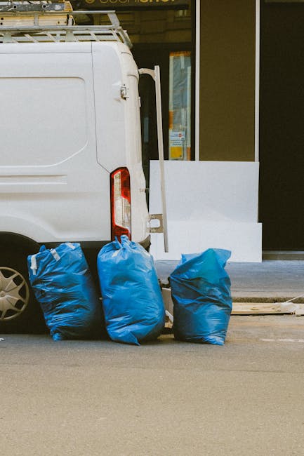 The image shows the rear section of a white commercial van parked on a street, with three large blue rubbish bags positioned on the pavement next to it. The bags appear to be made of heavy-duty plastic, with a slightly crinkled texture and some visible creases, indicating they are filled with waste. Behind the van, there is a building with a glass door or window, partially visible, and a light-colored step or ledge at the base of the entrance. In the background, part of a yellow-framed door or window is visible, suggesting an urban or commercial setting. The scene is well-lit, with natural daylight casting soft shadows on the street and the bags, emphasizing the contrast between the white van and the vibrant blue of the rubbish bags. The overall environment suggests a typical scenario where private waste collection, such as those offered by companies like wasteclearancewestkensington.co.uk, is used for on-site clearance of rubbish, avoiding delays associated with public or municipal waste disposal services.