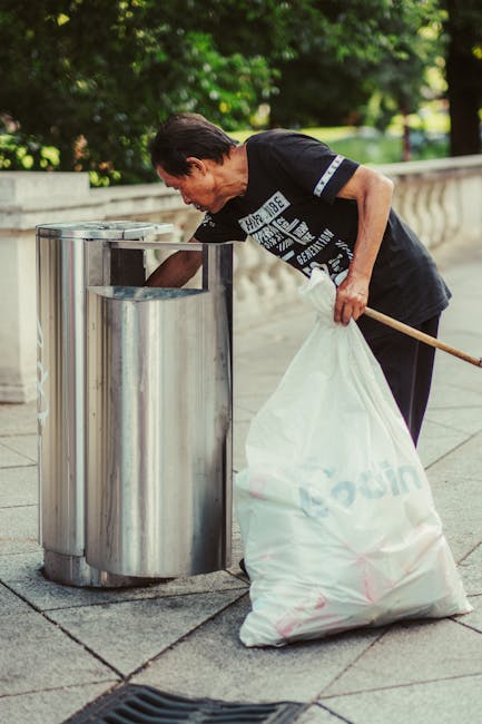 A middle-aged man with dark hair, dressed in a black T-shirt and dark trousers, is seen actively disposing of waste into a public recycling bin on a paved sidewalk. He is bending forward, holding a large white trash bag in his left hand, which appears to be filled with rubbish, and using his right hand to open the metal lid of the recycling container. The bin is cylindrical, made of stainless steel with a shiny finish, and has an open slot at the top for waste collection. The setting is outdoors, with a background of lush green trees and a stone balustrade along the sidewalk, indicating a park or urban residential area. The lighting suggests daytime with natural sunlight illuminating the scene, creating a clear and neutral atmosphere. This image illustrates responsible waste disposal and can relate to private rubbish collection or on-site clearance services sometimes employed in locations where independent waste handling is preferred over municipal collection, aligning with themes of rubbish removal and waste management solutions provided by [COMPANY_NAME].