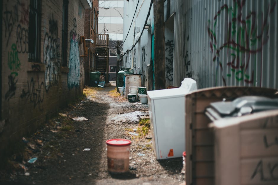 The image depicts a narrow alleyway between two brick and metal fence structures, both of which are covered in graffiti. Visible on the ground are patches of wet and muddy soil, with scattered debris and pieces of litter. Several large plastic containers and bin bags are placed along the sides, some with lids and others open, indicating they may contain waste materials. A small, red plastic bucket sits near the foreground, with other bins and recycling boxes positioned further back in the alley. The environment appears to be an urban backstreet or service access road, with a staircase and building features visible at the far end. The scene is lit with natural daylight, highlighting the textures of the graffiti, weathered surfaces, and waste materials. This setup suggests a location suited to private rubbish collection or on-site clearance services like those provided by waste clearance companies such as wasteclearancewestkensington.co.uk, for managing domestic or commercial waste outside of regular council pickups.