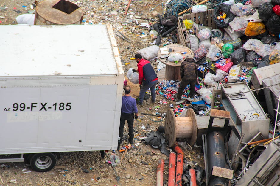 A middle-aged man with dark hair, dressed in a black T-shirt and dark trousers, is seen actively disposing of waste into a public recycling bin on a paved sidewalk. He is bending forward, holding a large white trash bag in his left hand, which appears to be filled with rubbish, and using his right hand to open the metal lid of the recycling container. The bin is cylindrical, made of stainless steel with a shiny finish, and has an open slot at the top for waste collection. The setting is outdoors, with a background of lush green trees and a stone balustrade along the sidewalk, indicating a park or urban residential area. The lighting suggests daytime with natural sunlight illuminating the scene, creating a clear and neutral atmosphere. This image illustrates responsible waste disposal and can relate to private rubbish collection or on-site clearance services sometimes employed in locations where independent waste handling is preferred over municipal collection, aligning with themes of rubbish removal and waste management solutions provided by [COMPANY_NAME].