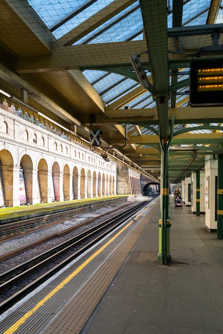 The image depicts an underground train station platform with multiple railway tracks extending into the distance. The platform surface is made of concrete, with a tactile paving strip along the edge for safety. Overhead, there is a partially enclosed, curved glass roof supported by green metal beams and columns, allowing natural light to illuminate the area. Along the platform, a few passengers are visible, including a person sitting on a bench and another walking with a small suitcase or bag. On the right side, there are rectangular white and green station signage and a small electronic display screen. The background shows an arched brick viaduct with multiple arches, a common feature in urban railway infrastructure. The station is clean and well-maintained, with an environment suitable for discreet rubbish collection or private waste management, aligning with independent collection practices that may occur on such platforms. The overall scene reflects typical elements of a metropolitan railway setting, connecting to broader themes of public transit and alternative waste handling adjacent to passenger service areas.