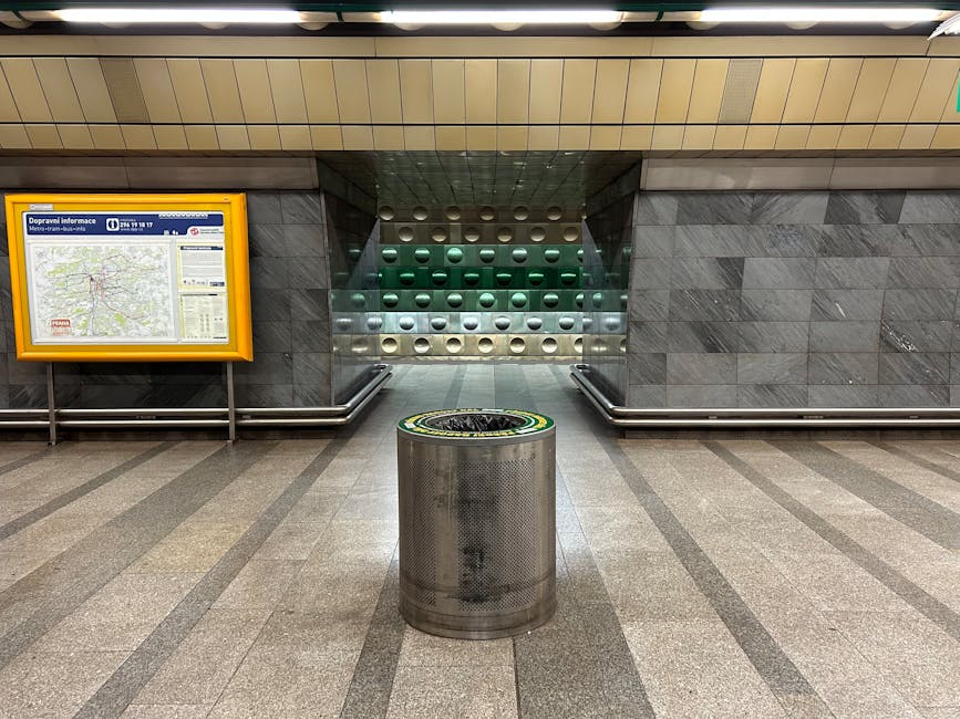 The image depicts an underground train station platform with multiple railway tracks extending into the distance. The platform surface is made of concrete, with a tactile paving strip along the edge for safety. Overhead, there is a partially enclosed, curved glass roof supported by green metal beams and columns, allowing natural light to illuminate the area. Along the platform, a few passengers are visible, including a person sitting on a bench and another walking with a small suitcase or bag. On the right side, there are rectangular white and green station signage and a small electronic display screen. The background shows an arched brick viaduct with multiple arches, a common feature in urban railway infrastructure. The station is clean and well-maintained, with an environment suitable for discreet rubbish collection or private waste management, aligning with independent collection practices that may occur on such platforms. The overall scene reflects typical elements of a metropolitan railway setting, connecting to broader themes of public transit and alternative waste handling adjacent to passenger service areas.